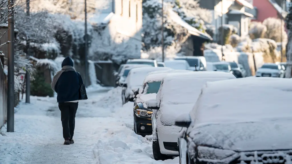 Eine-Person-geht-auf-einem-verschneiten-Gehweg-Am-Strassenrand-parken-mit-Schnee-bedeckte-Autos-Im-Tagesverlauf-des-Sonntags-herrscht-auflockernde-Bewoelkung-und-oft-Sonne-bei-Hoechstwerten-von-7-Grad