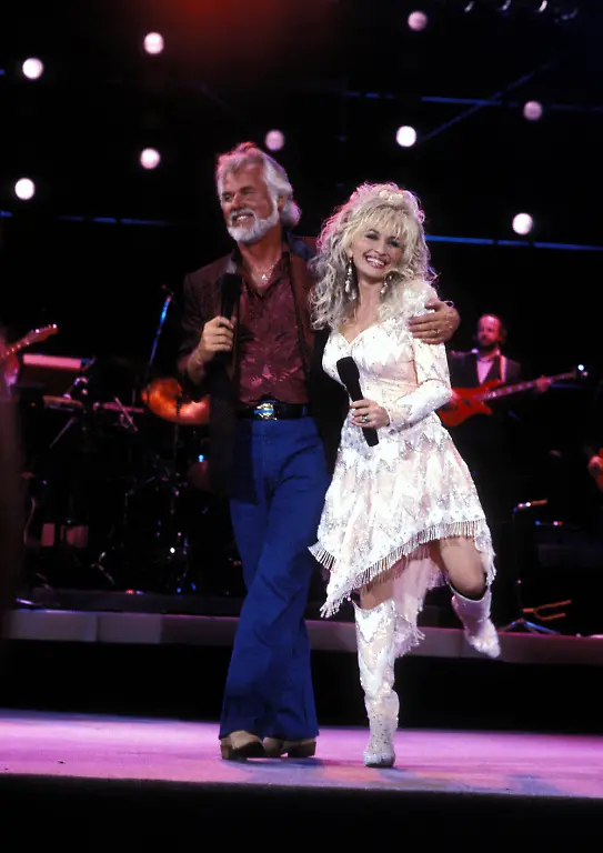 KENNY-ROGERS-August-21-1938-March-20-2020-was-an-American-singer-songwriter-actor-record-producer-and-entrepreneur-who-was-elected-to-the-Country-Music-Hall-of-Fame-in-2013-Rogers-was-particularly-popular-with-country-audiences-but-also-charted-more-than-120-hit-singles-across-various-music-genres-topped-the-country-and-pop-album-charts-for-more-than-200-individual-weeks-in-the-United-States-alone-He-sold-over-100-million-records-worldwide-during-his-lifetime-making-him-one-of-the-best-selling-music-artists-of-all-time-PICTURED-KENNY-ROGERS-and-DOLLY-PARTON-on-stage-in-1989-U-S