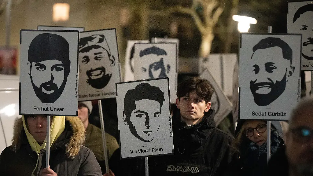 Demonstranten-halten-Plakate-mit-stilisierten-Bildern-der-Opfer-bei-einem-Gedenken-zum-Jahrestag-des-rassistischen-Anschlages-von-Hanau-am-damaligen-Tatort-in-Kesselstadt-Ein-43-jaehriger-Deutscher-hatte-am-19-Februar-2020-insgesamt-neun-Menschen-mit-Migrationshintergrund-ermordet