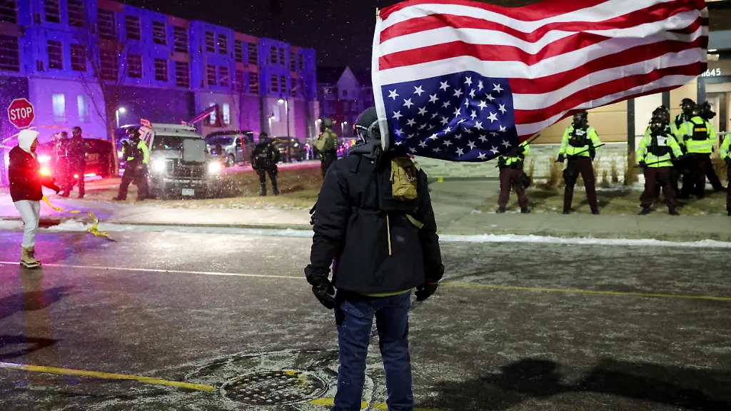 A-demonstrator-carrying-a-U-S-flag-walks-next-to-law-enforcement-officers-in-position-during-a-protest-outside-the-hotel-where-Greg-Bovino-who-has-been-removed-from-his-role-as-the-commander-at-large-for-the-U-S-Border-Patrol-is-reportedly-staying-in-Maple-Grove-Minnesota-U-S-January-26-2026-REUTERS-Shannon-Stapleton-REFILE-CORRECTING-LOCATION-FROM-MINNEAPOLIS-TO-MAPLE-GROVE