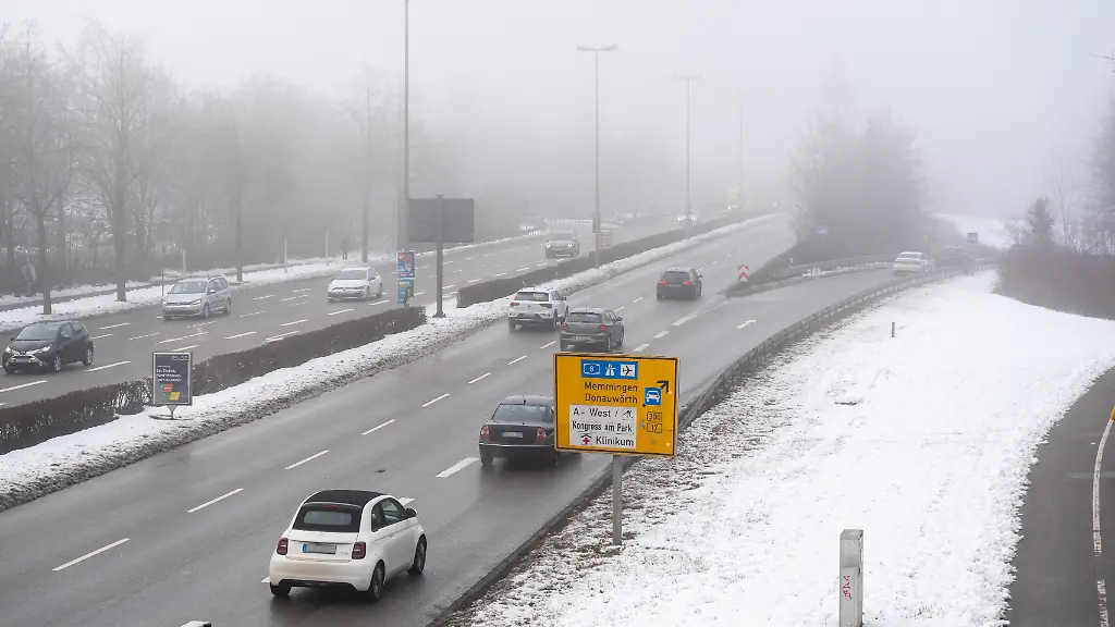 Staedtischer-Verkehr-auf-einer-breiten-Ausfallstrasse-im-Winter-Autos-fahren-bei-Nebel-und-Kaelte-in-Richtung-Innenstadt-waehrend-Schnee-und-Glaette-die-Aufmerksamkeit-der-Verkehrsteilnehmer-erhoehen