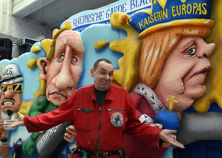 Der-Kuenstler-Jacques-Tilly-Karnevalswagenbauer-des-Duesseldorfer-Karnevalszugs-steht-am-06-02-2016-in-Duesseldorf-Nordrhein-Westfalen-in-der-Wagenbauhalle-mit-ausgebreiteten-Armen-vor-dem-Motivwagen-mit-Angela-Merkel-als-Kaiserin-Europas-r-und-Putin-mit-einer-Bombe-in-den-Haenden-Ganz-links-ein-Saudi-mit-einer-Zuendschnur-Ob-der-Wagen-allerdings-am-Rosenmontag-08-02-2016-auf-der-Strasse-zu-sehen-sein-wird-ist-fraglich-Dem-Rosenmontagszug-droht-aus-Sicherheitsgruenden-die-Absage-Die-Metreologen-rechnen-mit-Windstaerken-und-Sturmboeen