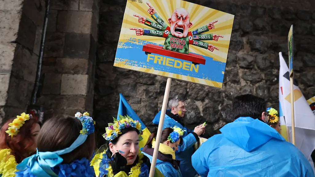 16-02-2026-Nordrhein-Westfalen-Koeln-Karnevalisten-des-Blau-Gelben-Kreuzes-tragen-ein-Schild-eines-frueheren-Satiremotivs-von-Jacques-Tilly-Der-Rosenmontag-am-Rhein-ist-der-Hoehepunkt-der-rheinischen-Karnevalssaison-mit-den-traditionellen-Umzuegen