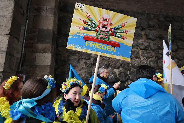 16-02-2026-Nordrhein-Westfalen-Koeln-Karnevalisten-des-Blau-Gelben-Kreuzes-tragen-ein-Schild-eines-frueheren-Satiremotivs-von-Jacques-Tilly-Der-Rosenmontag-am-Rhein-ist-der-Hoehepunkt-der-rheinischen-Karnevalssaison-mit-den-traditionellen-Umzuegen