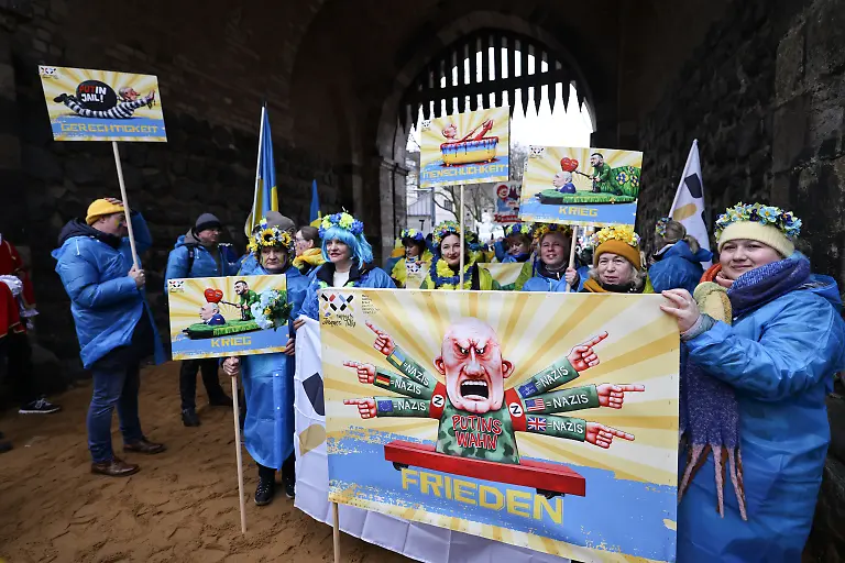 16-02-2026-Nordrhein-Westfalen-Koeln-Karnevalisten-des-Blau-Gelben-Kreuzes-tragen-ein-Schild-eines-frueheren-Satiremotivs-von-Jacques-Tilly-Der-Rosenmontag-am-Rhein-ist-der-Hoehepunkt-der-rheinischen-Karnevalssaison-mit-den-traditionellen-Umzuegen