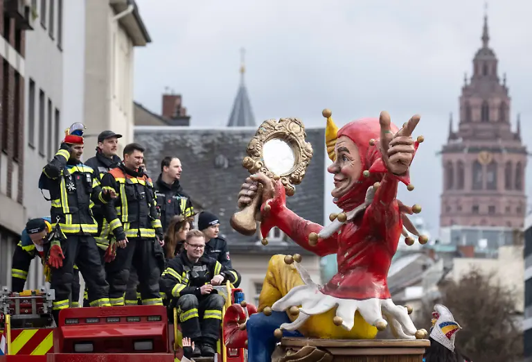 Ein-Motivwagen-mit-einer-ueberlebensgrossen-Narrenfigur-zieht-beim-Rosenmontagsumzug-durch-die-Stadt-Im-Hintergrund-ist-der-Mainzer-Dom-zu-sehen-waehrend-Einsatzkraefte-der-Feuerwehr-auf-einem-Fahrzeug-den-Zug-begleiten-Tausende-Zuschauer-verfolgen-den-Umzug-durch-das-Zentrum-der-Stadt-Der-naerrische-Lindwurm-umfasst-politische-Motivwagen-Schwellkoeppe-Musikzuege-Fahnentraeger-und-Garden-Die-Laenge-der-Zugstrecke-betraegt-ueber-sieben-Kilometer