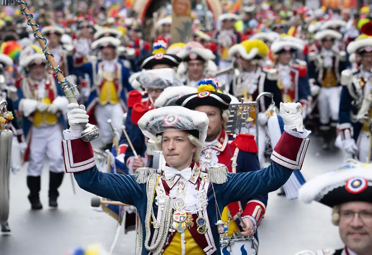 Teilnehmer-in-historischen-Uniformen-und-mit-Dreispitzhueten-marschieren-beim-Rosenmontagsumzug-durch-die-Stadt-Tausende-Zuschauer-verfolgen-den-Umzug-durch-das-Zentrum-der-Stadt-Der-naerrische-Lindwurm-umfasst-politische-Motivwagen-Schwellkoeppe-Musikzuege-Fahnentraeger-und-Garden-Die-Laenge-der-Zugstrecke-betraegt-ueber-sieben-Kilometer