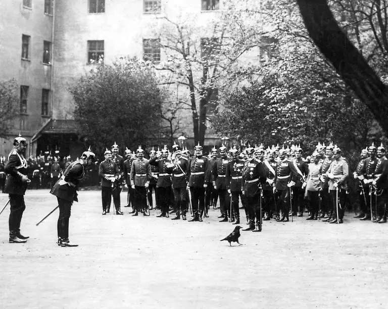 Undatierte-Aufnahme-Kaiser-Wilhem-II-vorgebaeugt-begruesst-auf-Besuch-beim-Kaiser-Alexander-Garde-Grenadier-Regiment-Nr-1-in-der-Kaserne-in-der-Prinz-Friedrich-Karl-Strasse-heute-Geschwister-Scholl-Strasse-in-Berlin-Mitte-einen-zahmen-Kolkraben-das-Maskottchen-des-Regiments-Dahinter-beobachten-Offiziere-des-Regiments-die-Szenerie-In-der-Mitte-steht-ein-russischer-Offizier-mit-Pelzmuetze-und-russischer-Uniform-Zar-Nikolaus-II-von-Russland-war-bis-1914-offiziell-Kommandeur-des-preussischen-Regiments-Aufnahmedatum-01-01-1900-31-12
