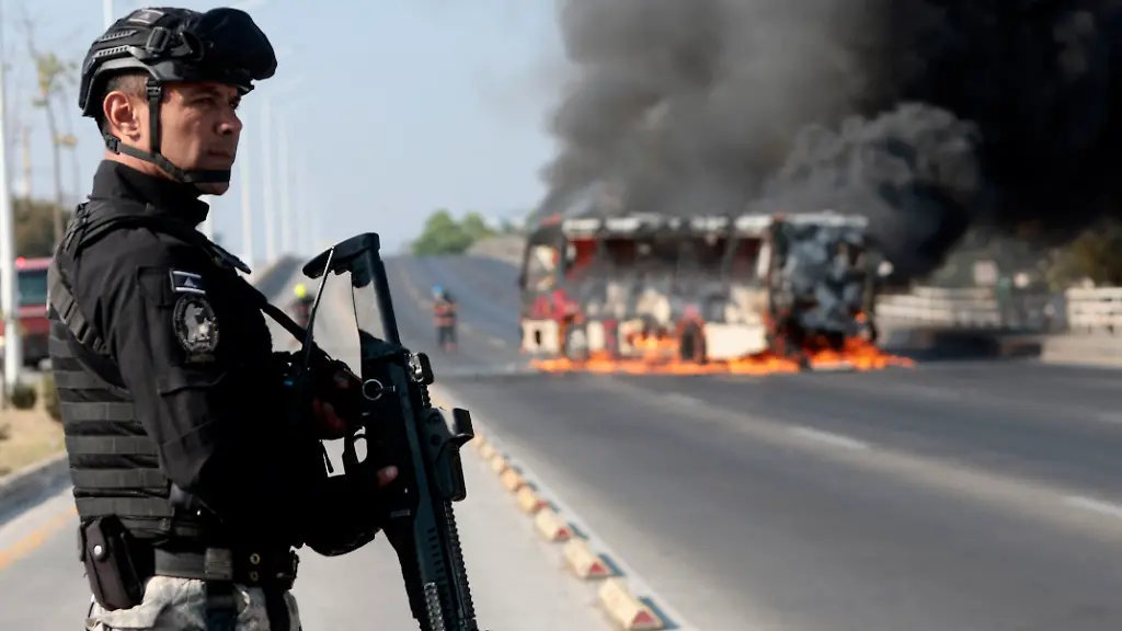 A-member-of-the-Prosecutor-s-Office-stands-guard-near-a-burning-bus-at-one-of-the-main-avenues-after-it-was-set-on-fire-by-organised-crime-groups-in-response-to-an-operation-in-Jalisco-to-arrest-a-high-priority-security-target-in-Zapopan-state-of-Jalisco-Mexico-on-February-22-2026-Armed-civilians-blocked-several-roads-in-the-state-of-Jalisco-in-western-Mexico-following-an-operation-by-federal-forces-in-the-town-of-Tapalpa-local-authorities-reported-Jalisco-which-will-host-four-matches-of-the-upcoming-2026-World-Cup-is-home-to-the-powerful-Jalisco-New-Generation-Cartel-CJNG-and-has-been-rocked-by-several-episodes-of-violence-in-recent-years