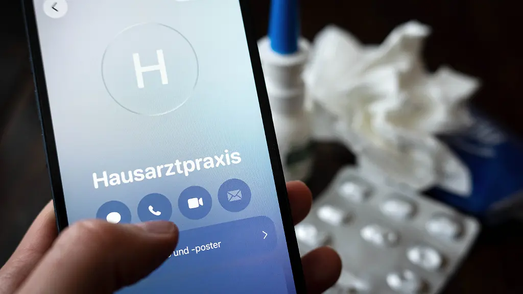 ILLUSTRATION-18-02-2026-Bremen-Eine-Person-haelt-ein-Smartphone-in-der-Hand-und-ruft-eine-Hausarztpraxis-an-Im-Hintergrund-liegen-ein-Blister-mit-Tabletten-ein-Nasenspray-und-ein-Taschentuch-auf-einem-Tisch