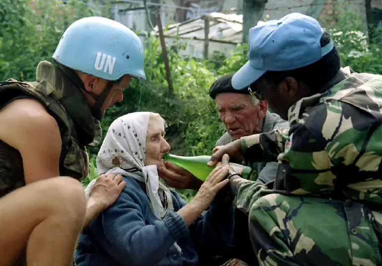 A-Kenyan-UN-peacekeeper-with-his-Dutch-comrade-gives-water-to-an-elderly-Bosnian-Moslem-woman-refugee-from-Srebrenica-as-she-waits-to-be-transported-from-the-eastern-Bosnian-village-of-Potocari-to-Moslem-held-Kladanj-near-Olovo-July-13-1995-On-July-11-1995-towards-the-end-of-Bosnia-s-1992-95-war-Bosnian-Serb-forces-swept-into-the-eastern-Srebrenica-enclave-a-U-N-designated-safe-heaven-There-they-took-8-000-Muslim-men-and-boys-and-executed-them-in-the-days-that-followed-dumping-their-bodies-into-pits-in-the-surrounding-forests