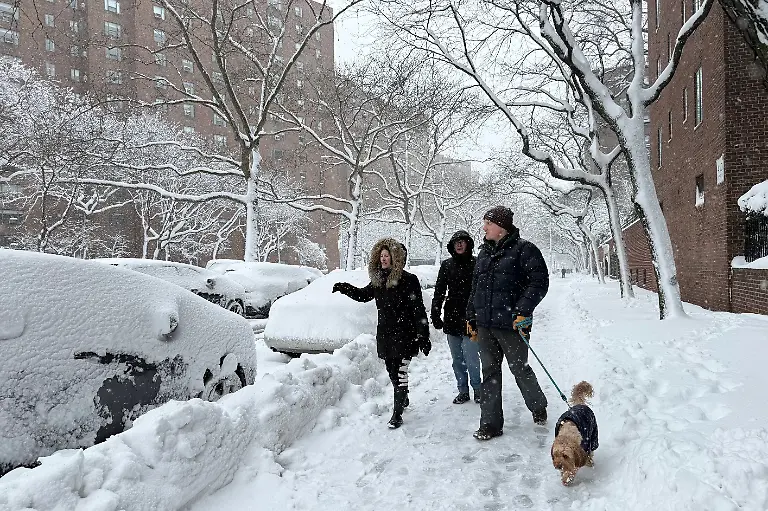 People-walk-with-a-dog-alongside-snow-laden-parked-cars-in-a-snowstorm-Monday-Feb-23-2026-in-New-York