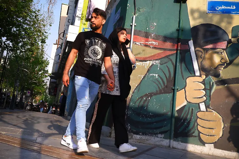 April-14-2024-Tehran-Iran-An-Iranian-young-couple-walks-next-to-a-wall-painting-of-Iranian-Revolutionary-Guard-Corps-IRGC-soldiers-at-Palestine-Square-in-downtown-Tehran-Israel-on-Sunday-hailed-its-air-defenses-in-the-face-of-an-unprecedented-attack-by-Iran-saying-the-systems-thwarted-99-of-the-more-than-300-drones-and-missiles-launched-toward-its-territory