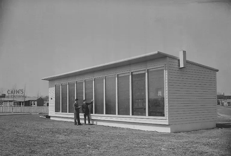 Paul-Witherell-research-director-at-Massachusetts-Institute-of-Technology-right-reviews-a-thermometer-s-measure-of-outside-air-temperature-on-the-south-wall-of-the-experimental-building-in-Cambridge-Mass-March-28-1947-The-glass-sections-are-double-glass-windows-behind-which-are-arranged-various-types-of-heat-storing-units-including-sealed-cells-of-chemicals-The-other-individual-is-unidentified-M-I-T-constructed-a-small-building-to-experiment-with-collecting-and-utilizing-the-sun-s-energy-for-heat-Poorly-insulated-panels-are-inserted-to-achieve-a-controlled-heat-loss-and-walls-adjoining-cubicles-are-well-insulated-to-permit-simultaneous-but-independent-experiments-Heat-is-collected-and-stored-in-walls-forming-a-part-of-the-house-s-south-wall-Walls-may-be-either-massive-enough-to-store-heat-over-short-periods-by-a-moderate-rise-in-temperature-or-they-may-contain-a-chemical-compound-which-melts-at-the-normal-temperature-of-domestic-radiant-heating-panels-and-which-stores-heat-by-its-melting-To-prevent-heat-loss-to-the-outdoors-and-permit-absorption-of-solar-energy-the-walls-are-covered-with-double-glass-panes-and-may-also-be-outfitted-with-shades-that-automatically-draw-when-the-sun-is-not-shining-Dr-Maria-Telkes-research-associate-at-M-I-T-s-Department-of-Metallurgy-developed-the-method-of-storing-heat-in-cells-containing-chemicals-that-is-being-studied-in-this-experimental-building-In-this-instance-the-chemical-is-stored-in-ordinary-tin-cans-which-are-sealed-before-being-placed-in-the-building-wall-Because-the-project-is-purely-experimental-the-particular-construction-used-is-in-no-sense-typical-of-a-dwelling-built-to-utilize-the-principle-being-studied