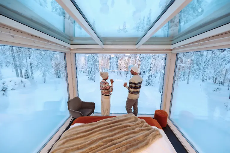 Zwei-Personen-geniessen-warme-Getraenke-in-einem-modernen-Glas-Iglu-umgeben-von-einer-schneebedeckten-Waldlandschaft-in-Akaslompolo-Lappland-Eine-gemuetliche-Atmosphaere-laedt-zur-Entspannung-und-Verbindung-mit-der-Natur-ein