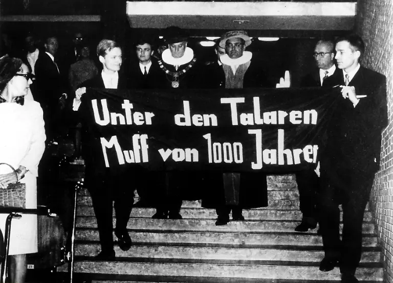 Deutschland-im-Herbst-1967-Feier-des-Rektorenwechsels-an-der-Universitaet-Hamburg-Die-Studenten-der-Universitaet-Hamburg-Detlev-Albers-li-und-Gert-Hinnerk-Behlmer-demonstrieren-im-Audimax-gegen-die-Ernennung-des-Universitaetsrektors-mit-dem-Spruchband-Unter-den-Talaren-Muff-von-1000-Jahren-dahinter-links-der-ehemalige-Rektor-Prof-Schoefer-rechts-der-neue-Prof-Ehrlicher-09-11