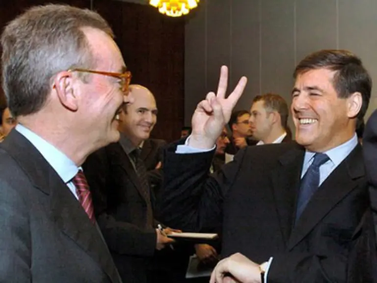 Breit-laechelnd-hebt-der-Bank-Boss-die-rechte-Hand-und-formt-aus-Zeige-und-Mittelfinger-ein-V