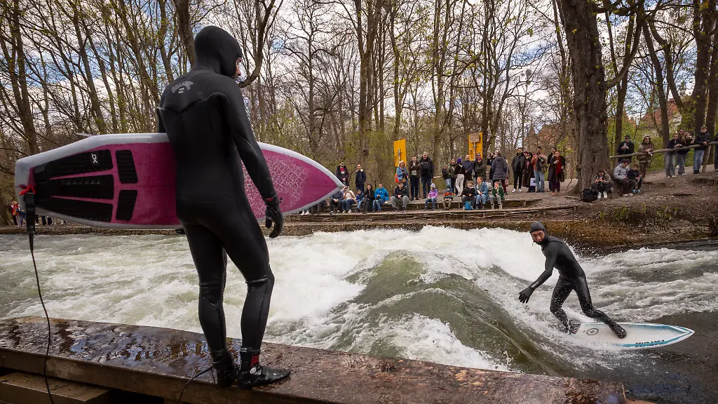 Ein-Mann-mit-Surfbrett-schaut-einen-Surfer-zu-der-mit-seinem-Board-auf-der-provisorischen-Eisbachwelle-im-Englischen-Garten-reitet-Die-Welle-verschwand-im-vergangenen-Herbst-und-hat-sich-seitdem-nicht-mehr-stabil-aufgebaut-Es-gab-inzwischen-mehrere-Versuche-sie-wieder-verlaesslich-surfbar-zu-machen-Die-Welle-ist-normalerweise-zu-jeder-Jahres-und-Tageszeit-ein-wahrer-Hotspot-fuer-Touristen-und-Surfer-aus-der-ganzen-Welt