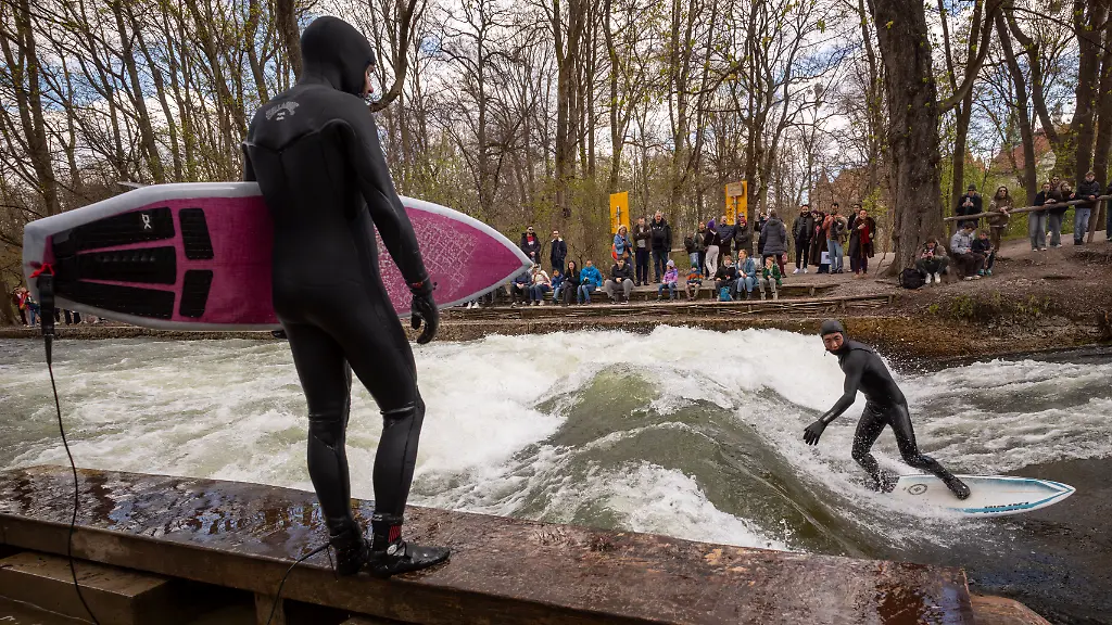 Ein-Mann-mit-Surfbrett-schaut-einen-Surfer-zu-der-mit-seinem-Board-auf-der-provisorischen-Eisbachwelle-im-Englischen-Garten-reitet-Die-Welle-verschwand-im-vergangenen-Herbst-und-hat-sich-seitdem-nicht-mehr-stabil-aufgebaut-Es-gab-inzwischen-mehrere-Versuche-sie-wieder-verlaesslich-surfbar-zu-machen-Die-Welle-ist-normalerweise-zu-jeder-Jahres-und-Tageszeit-ein-wahrer-Hotspot-fuer-Touristen-und-Surfer-aus-der-ganzen-Welt
