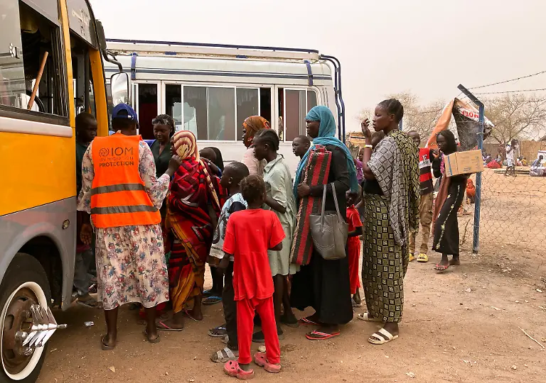 Eine-UN-Mitarbeiterin-l-steht-mit-wartenden-Fluechtlingen-an-einem-Bus-der-Frauen-und-Kinder-aus-dem-Sudan-vom-Grenzuebergang-Joda-in-ein-Transitlager-in-der-Grenzstadt-Renk-im-Suedsudan-bringen-soll-Der-Machtkampf-im-Sudan-der-seit-Mitte-April-2023-andauert-hat-die-nach-UN-Angaben-weltweit-groesste-Fluechtlingskrise-mit-mehr-als-acht-Millionen-Gefluechteten-ausgeloest-Waehrend-viele-dieser-Menschen-innerhalb-des-Sudans-Sicherheit-vor-den-Kaempfen-suchen-flohen-andere-in-die-Nachbarlaender-Suedsudan-oder-Tschad