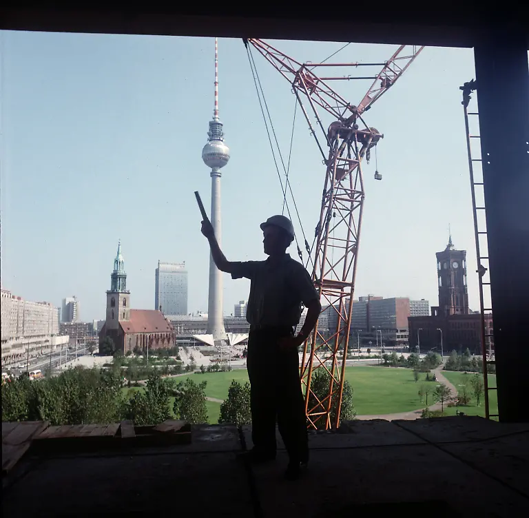 Ein-Bauingenieur-steht-als-Silhouette-in-einer-Oeffnung-einer-Rohbaufassade-auf-der-Baustelle-des-Palast-der-Republik-in-Berlin-Ost-im-Hintergrund-die-Neubauten-an-der-Karl-Liebknecht-Strasse-l-r-die-historische-Marienkirche-das-Hochhaushotel-Stadt-Berlin-der-Fernsehturm-Neubauten-an-der-Rathauspassage-und-das-Rote-Rathaus-undatiertes-Foto-aus-dem-Jahr-1974-In-den-1970er-Jahren-begann-die-architektonische-Umgestaltung-von-Berlin-Ost-zur-sozialistischen-Hauptstadt