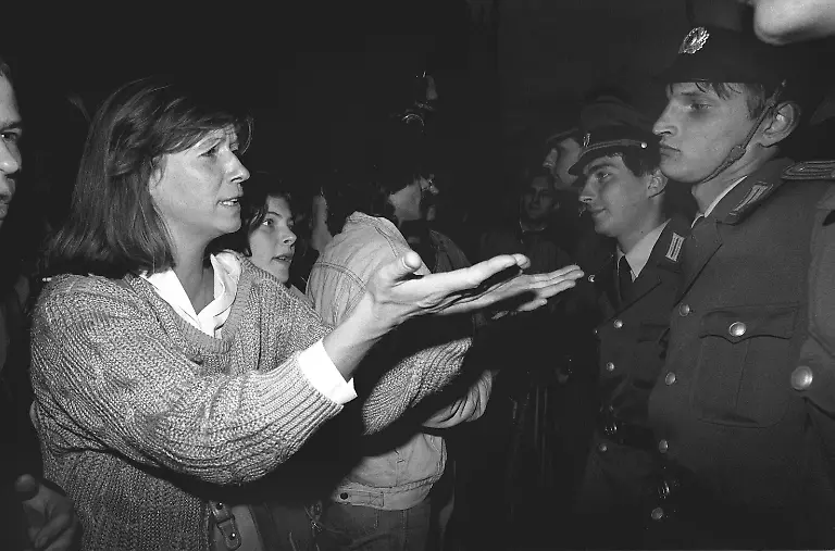 Mit-einer-Geste-versucht-eine-Frau-am-7-Oktober-1989-bei-einer-Demonstration-in-Ostberlin-Beamte-in-einer-Polizeikette-von-ihrer-Friedfertigkeit-zu-ueberzeugen-Am-7-Oktober-1989-kam-es-wie-in-anderen-ostdeutschen-Staedten-auch-in-Ostberlin-zu-Demonstrationen-gegen-die-Machthaber-in-der-DDR-Am-Rande-der-offiziellen-Feierlichkeiten-zum-40-Jahrestag-der-Republikgruendung-bildete-sich-auf-dem-Alexanderplatz-ein-Protestzug-mit-rund-1000-meist-jugendlichen-Teilnehmern-der-zu-dem-von-Sicherheitskraeften-abgesperrten-Palast-der-Republik-marschierte-Im-Verlauf-der-Demonstration-gingen-Volkspolizei-und-zivile-Beamte-der-Staatssicherheit-zum-Teil-massiv-gegen-Demonstranten-und-Journalisten-vor