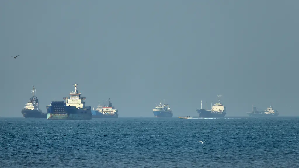 Frachtschiffe-darunter-Massengutfrachter-und-Stueckgutfrachter-liegen-vor-der-Kueste-vor-Anker-waehrend-im-Vordergrund-ein-kleines-Motorboot-in-der-Strasse-von-Hormuz-vor-Bandar-Abbas-Iran-vorbeifaehrt