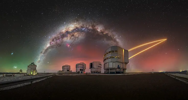 This-scene-was-captured-at-the-Very-Large-Telescope-VLT-on-Cerro-Paranal-in-Chile-s-Atacama-Desert-under-exceptionally-pure-observing-conditions-where-the-boundary-between-Earth-and-sky-feels-almost-seamless-The-Milky-Way-stretches-across-the-sky-with-its-dense-central-bulge-rising-prominently-overhead-To-the-left-the-Magellanic-Clouds-two-neighboring-satellite-galaxies-are-visible-to-the-naked-eye-from-the-Southern-Hemisphere-Subtle-airglow-adds-another-layer-to-the-scene-a-natural-emission-produced-by-chemical-reactions-in-the-upper-atmosphere-at-altitudes-of-80-to-100-kilometers-In-this-image-it-reveals-a-range-of-colors-with-green-tones-on-the-left-and-warmer-reddish-hues-toward-the-right-In-the-foreground-stands-the-VLT-operated-by-the-European-Southern-Observatory-Situated-at-2-635-meters-above-sea-level-the-site-benefits-from-extremely-dry-conditions-stable-atmospheric-layers-and-minimal-light-pollution-resulting-in-more-than-300-clear-nights-each-year-The-observatory-consists-of-four-primary-telescopes-each-equipped-with-8-2-meter-mirrors-One-of-them-UT4-Yepun-projects-four-sodium-laser-beams-into-the-sky-creating-artificial-guide-stars-at-an-altitude-of-approximately-90-kilometers-These-reference-points-enable-adaptive-optics-systems-to-correct-for-atmospheric-distortion-in-real-time-significantly-enhancing-the-clarity-and-precision-of-astronomical-observations