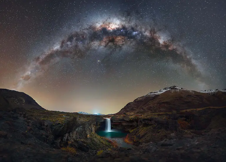 This-image-was-captured-during-a-night-at-Salto-del-Agrio-in-the-province-of-Neuquen-Argentina-It-s-a-place-that-invites-reflection-on-the-immense-sequence-of-events-both-on-Earth-and-in-the-sky-that-shaped-this-landscape-The-45-meter-waterfall-plunges-into-a-canyon-carved-by-ancient-lava-flows-from-the-Copahue-volcano-Surrounded-by-basalt-the-scene-is-enriched-by-the-vivid-colors-left-behind-by-minerals-particularly-iron-and-sulfur-carried-by-the-Agrio-River-Images-like-this-are-carefully-planned-and-patiently-awaited-yet-they-always-carry-an-element-of-uncertainty-Weather-is-often-the-greatest-challenge-My-first-attempt-to-reach-this-location-was-stopped-by-heavy-snowfall-and-on-this-occasion-the-cold-and-strong-winds-once-again-put-the-outcome-in-doubt-Despite-the-harsh-conditions-I-waited-for-the-precise-moment-when-the-Milky-Way-aligned-above-the-waterfall-forming-an-arch-suspended-over-this-ancient-terrain-Working-with-the-tripod-close-to-the-ground-and-repeating-exposures-I-was-finally-able-to-capture-the-scene-I-had-envisioned-for-so-long