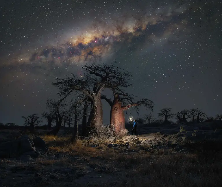 I-spent-ten-days-traveling-through-Botswana-living-out-of-my-car-and-moving-through-remote-desert-landscapes-in-complete-isolation-focused-entirely-on-photographing-the-night-sky-This-location-became-the-highlight-of-the-trip-a-small-ancient-island-rising-from-the-middle-of-a-vast-salt-pan-Reaching-it-however-was-far-from-straightforward-After-struggling-to-find-the-route-and-nearly-giving-up-I-only-managed-to-get-there-by-chance-spotting-two-Jeeps-heading-in-the-opposite-direction-and-deciding-to-turn-around-and-follow-them-After-three-intense-hours-navigating-the-terrain-I-finally-found-myself-driving-across-the-open-salt-pan-leaving-a-trail-of-white-dust-behind-The-island-itself-was-covered-with-baobab-trees-scattered-in-small-groups-or-standing-alone-each-one-offering-a-striking-subject-under-the-night-sky-Many-were-naturally-positioned-beneath-the-Milky-Way-core-early-in-the-night-while-others-lent-themselves-to-different-compositions-including-star-trails-This-particular-group-stood-out-to-me-the-most-Alone-in-the-darkness-I-took-my-time-shaping-the-scene-using-a-torch-to-carefully-light-paint-the-foreground-The-final-image-is-a-blend-of-one-exposure-for-the-landscape-eleven-for-the-light-painting-and-a-stack-of-four-tracked-exposures-for-the-sky-combined-to-balance-detail-and-atmosphere