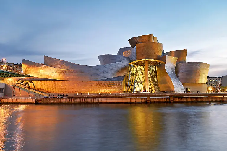 Visitors-admire-the-Guggenheim-Museum-in-Bilbao-as-it-lights-up-at-night-showcasing-its-unique-design-along-the-river
