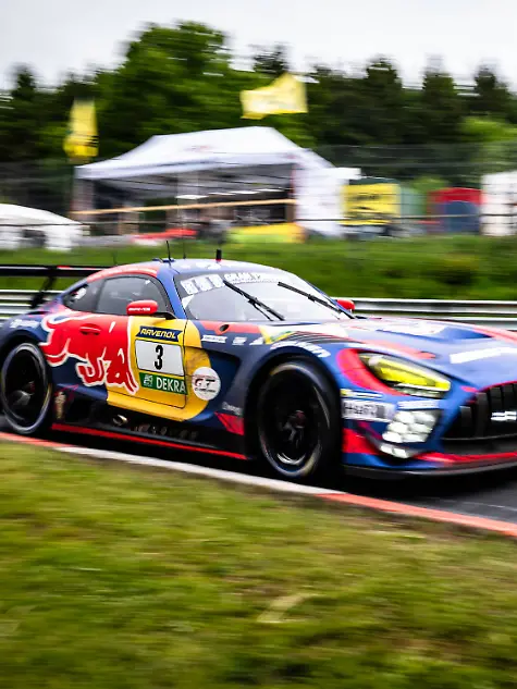Der-Mercedes-AMG-GT3-mit-der-Startnummer-3-von-Mercedes-AMG-Team-Verstappen-Racing-durchfaehrt-mit-hoher-Geschwindigkeit-eine-Kurve-auf-dem-Nuerburgring-Das-Fahrzeug-traegt-das-markante-Red-Bull-Livery-in-Blau-Rot-und-Gelb-mit-RAVENOL-und-DEKRA-Sponsoring-Die-Panning-Aufnahme-betont-die-Dynamik-des-Rennwagens-waehrend-der-Hintergrund-mit-Boxenanlagen-und-Baeumen-verschwimmt