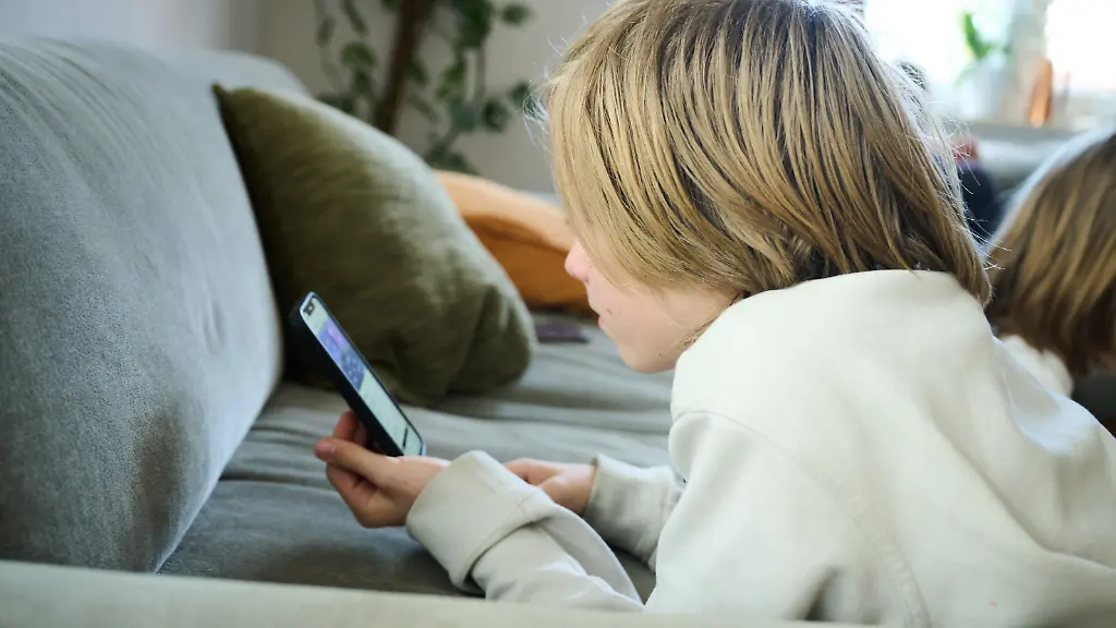 ARCHIV-21-04-2025-Bayern-Roth-Ein-Junge-liegt-auf-einem-Sofa-und-blickt-auf-sein-Smartphone-gestellte-Szene