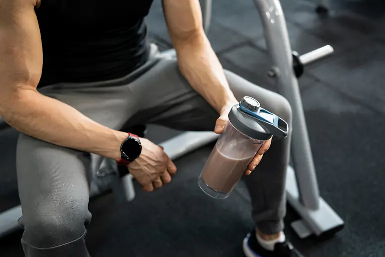 Muscular-man-resting-on-gym-machine-holding-shaker-bottle-with-protein-drink-after-training-session-Recovery-after-workout