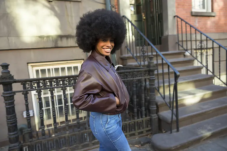 Junge-Afroamerikanerin-laechelt-waehrend-sie-mit-Afro-Frisur-und-brauner-Bomberjacke-vor-einem-klassischen-Brownstone-Wohnhaus-und-einem-schmiedeeisernen-Gelaender-in-Manhattan-posiert-Modellfreigabe-vorhanden