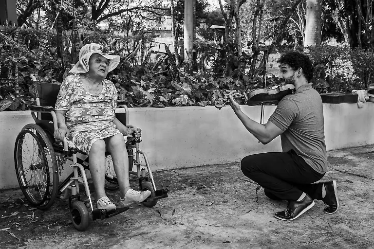 From-the-Violin-to-the-Heart-This-elderly-woman-was-taken-in-by-a-long-term-care-institution-for-older-adults-Lar-dos-Velinhos-de-Campinas-She-had-been-living-with-her-brother-in-a-shack-in-extremely-precarious-conditions-She-arrived-severely-ill-malnourished-suffering-from-dementia-Her-physical-and-mental-health-had-been-deeply-affected-by-a-lifetime-of-hardship-neglect-and-the-indifference-of-her-own-family-Within-the-institution-beyond-receiving-basic-healthcare-she-was-given-the-opportunity-to-age-with-dignity-She-now-takes-part-in-recreational-activities-designed-for-older-adults-occupational-therapy-workshops-and-daily-interaction-with-volunteers-each-offering-a-portion-of-their-time-care-and-personal-skills-A-young-conductor-and-violinist-volunteering-out-of-deep-sensitivity-to-the-challenges-faced-by-society-especially-older-people-in-situations-of-social-vulnerability-opened-his-heart-Through-music-he-offered-her-his-very-best-sending-a-message-directly-to-her-heart-As-he-played-her-expression-slowly-transformed-from-one-marked-by-exhaustion-and-the-weight-of-life-to-one-of-serenity-and-gentleness-In-a-deeply-moving-gesture-he-knelt-before-her-while-playing-as-if-offering-a-serenade-perhaps-the-first-she-had-ever-received-In-that-moment-his-act-seemed-to-say-From-my-youth-with-so-many-paths-still-to-walk-toward-aging-and-wisdom-I-offer-you-my-deepest-respect-love-tenderness-and-reverence-for-your-life