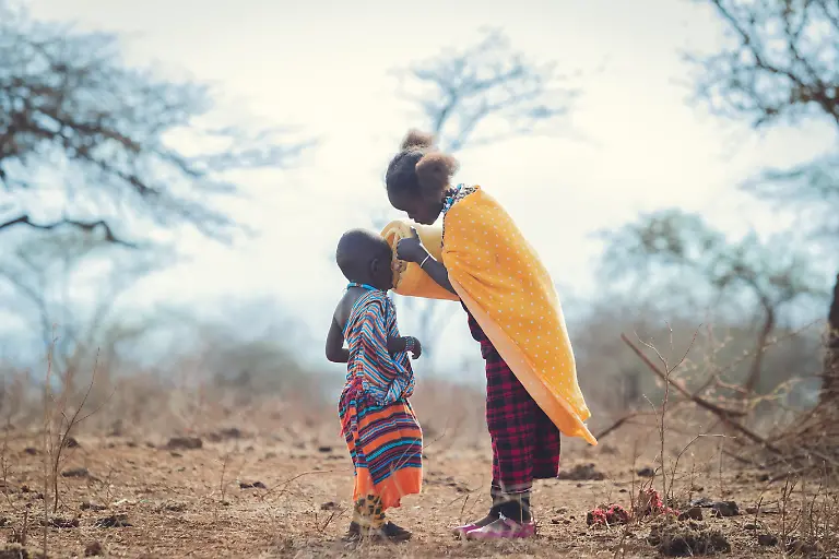 In-the-dry-open-landscape-where-the-earth-stretches-out-in-shades-of-brown-and-the-trees-stand-thin-against-the-sky-a-quiet-moment-of-tenderness-unfolds-Two-children-stand-close-wrapped-in-vibrant-traditional-fabrics-that-brighten-the-muted-surroundings-The-elder-child-gently-lifts-a-small-container-helping-the-younger-one-drink-her-posture-soft-patient-and-full-of-care-It-is-a-simple-act-yet-in-this-environment-it-speaks-volumes-Amid-the-dust-the-heat-and-the-challenges-of-everyday-life-this-moment-becomes-a-symbol-of-resilience-and-shared-strength-The-love-between-them-is-unspoken-but-unmistakable-a-bond-shaped-by-responsibility-trust-and-the-instinct-to-protect-Even-in-a-place-where-resources-are-limited-compassion-is-abundant-Their-brightly-colored-clothing-contrasts-beautifully-with-the-dry-earth-reminding-us-that-hope-too-can-stand-out-even-in-the-harshest-conditions-As-the-younger-child-receives-the-sip-their-posture-reflects-comfort-and-reliance-knowing-they-are-cared-for-And-in-the-elder-child-s-gentle-gesture-is-a-quiet-form-of-leadership-one-that-doesn-t-require-words-only-kindness-In-this-brief-exchange-the-landscape-is-transformed-It-becomes-a-reminder-that-humanity-s-greatest-strength-is-often-found-in-its-smallest-most-heartfelt-moments