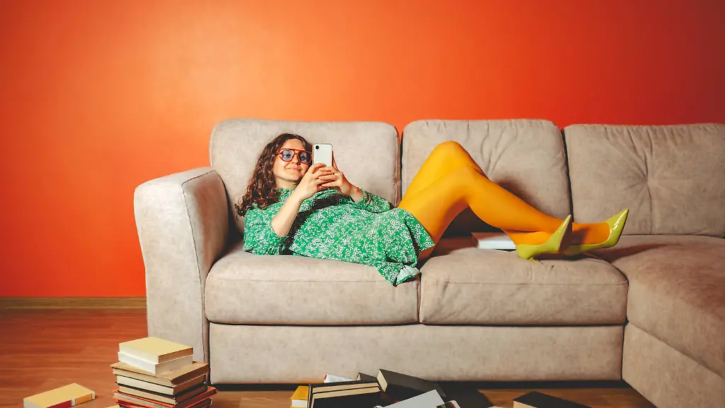 Person-lying-on-sofa-distracted-by-smartphone-surrounded-by-books-model-released-Symbolfoto-AAF00424