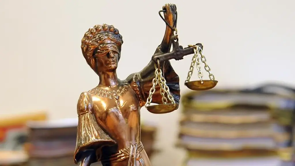 Eine-goldfarbene-Justitia-Figur-steht-vor-Aktenbergen-die-sich-auf-einem-Tisch-stapeln
