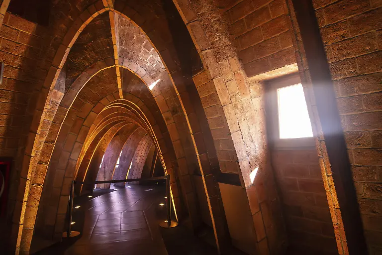 Warmes-sonnenlicht-stroemt-durch-ein-fenster-und-beleuchtet-die-einzigartigen-ziegelboegen-im-dachgeschoss-der-casa-mila-einem-beruehmten-architektonischen-wahrzeichen-in-barcelona-spanien