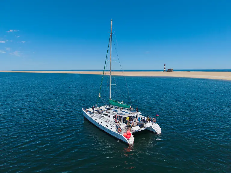 Luftaufnahme-des-Segelkatamarans-Silverskye-in-einer-Lagune-auf-dessen-Deck-Menschen-Austern-und-Champagner-geniessen-mit-einer-Sandbank-und-einem-Leuchtturm-in-der-Ferne-nahe-Walvis-Bay-Erongo-Namibia