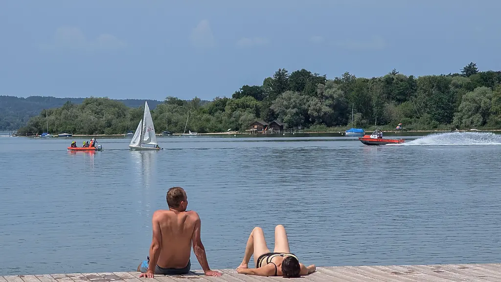 A-man-and-a-woman-sunbathe-on-a-wooden-pier-as-a-sailboat-and-motorboats-pass-on-Lake-Ammersee-in-Herrsching-am-Ammersee-Upper-Bavaria-Bavaria-Germany-on-May-31-2026-Lake-Ammersee-is-part-of-the-Land-of-Five-Lakes-Fuenfseenland-where-warm-weather-attracts-visitors-for-swimming-boating-sailing-and-lakeside-recreation