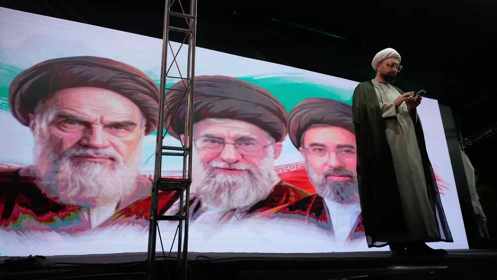 Ein-Geistlicher-ueberprueft-sein-Handy-auf-der-Buehne-vor-einem-Bildschirm-mit-Portraets-des-verstorbenen-iranischen-Revolutionsgruenders-Ayatollah-Khomeini-l-r-des-verstorbenen-Obersten-Fuehrers-Ayatollah-Ali-Khamenei-und-des-Obersten-Fuehrers-Ayatollah-Mojtaba-Khamenei-waehrend-einer-regierungsfreundlichen-Versammlung-in-Teheran-Iran