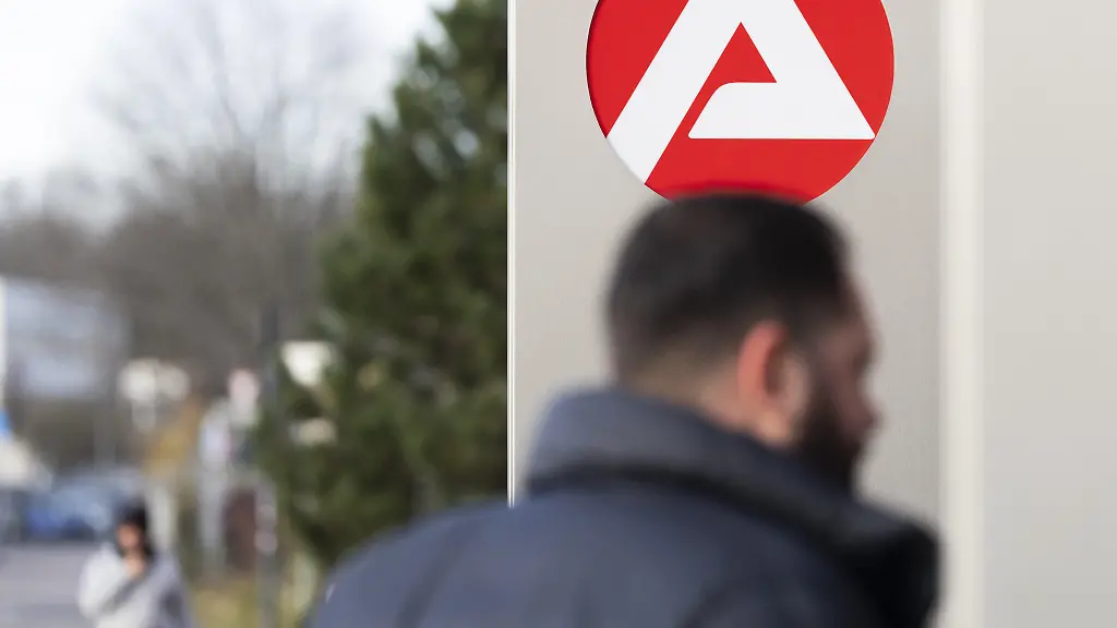 Stele-mit-dem-Zeichen-Logo-der-Agentur-fuer-Arbeit-vor-dem-Gebaeude-der-Agentur-fuer-Arbeit-unscharfe-Personen-auf-dem-Weg-zur-Agentur-Koeln-Nordrhein-Westfalen-Deutschland