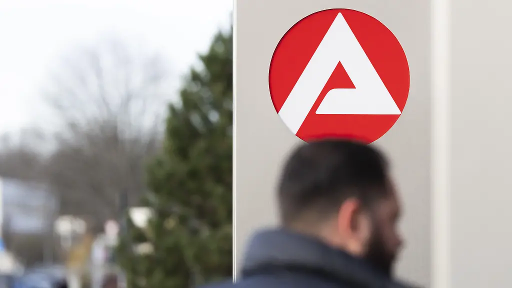 Stele-mit-dem-Zeichen-Logo-der-Agentur-fuer-Arbeit-vor-dem-Gebaeude-der-Agentur-fuer-Arbeit-unscharfe-Personen-auf-dem-Weg-zur-Agentur-Koeln-Nordrhein-Westfalen-Deutschland