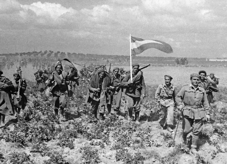 Aufnahme-nichtregulaerer-nationalspanischer-Truppen-beim-Marsch-ueber-ein-Feld-in-der-spanischen-Meseta-am-28-Maerz-1939-in-den-letzten-Tagen-des-Krieges-Die-Soldaten-tragen-ihr-gesamtes-Marschgepaeck-und-haben-nationalspanische-Fahnen-rot-gelb-rot-dabei-Im-Vordergrund-geht-ein-Leutnant-mit-gehstock-und-Zigarette-an-der-Spitze-Im-Hintegrund-sind-Olivenhaine-zu-sehen