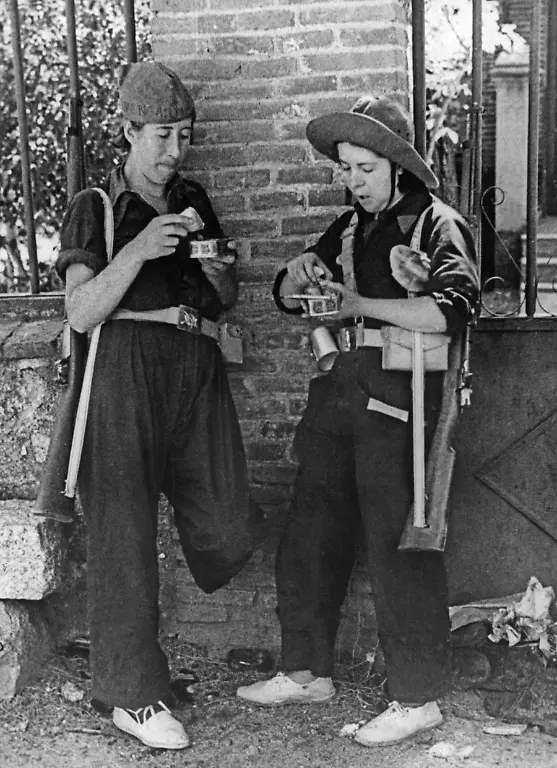 Aufnahme-zweier-republikanischer-Kaempferinnen-einer-Arbeitermiliz-in-der-Naehe-der-Front-in-der-Sierra-Guadarrama-noerdlich-von-Madrid-im-August-1936-kurz-nach-Ausbruch-des-Spanischen-Buergerkriegs-beim-Mittagessen-Die-beiden-Frauen-die-Koppeln-tragen-sind-mit-Karabinern-bewaffnet-Ihre-Mahlzeit-besteht-aus-Dosenfisch-Sie-tragen-Alpargatas-leichte-Baumwollschuhe-mit-einer-Sohle-aus-geflochtenem-Hanf-Die-beiden-lehnen-an-einer-Mauer-zu-einem-Vorgarten