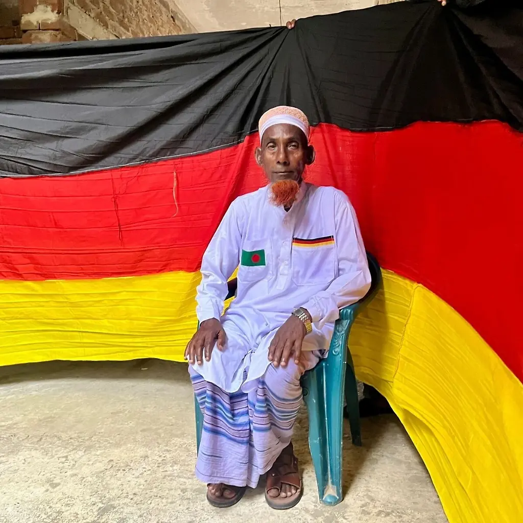 Der-77-jaehrige-bangladeschische-Bauer-Amjad-Hossain-posiert-mit-der-deutschen-Flagge-die-er-zu-Hause-genaeht-hat-Er-entrollte-die-7-5-Kilometer-lange-Flagge-am-10-Juni-auf-dem-Pausenplatz-einer-oertlichen-Schule-einen-Tag-vor-Beginn-der-FIFA-Weltmeisterschaft-2026