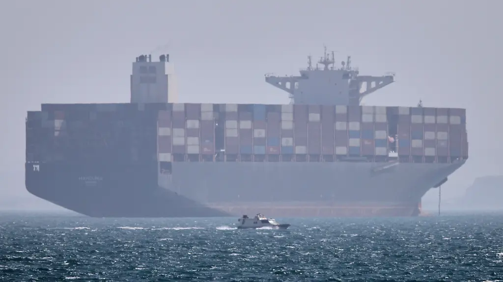 ARCHIV-02-05-2026-Iran-Bandar-Abbas-Ein-Containerschiff-liegt-vor-Anker-waehrend-im-Vordergrund-ein-kleines-Motorboot-in-der-Strasse-von-Hormus-vor-vorbeifaehrt