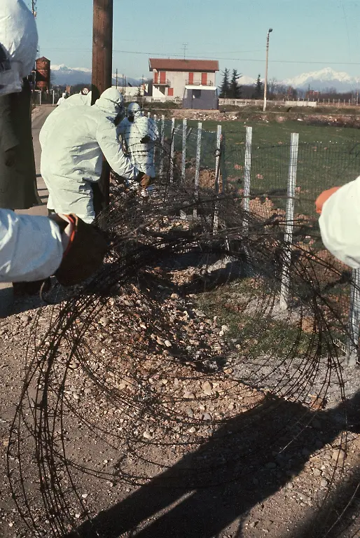 Arbeiter-in-Schutzanzuegen-zaeunen-mit-Dioxin-verseuchtes-Gelaende-im-Mailaender-Vorort-Seveso-mit-Stacheldraht-ein-aufgenommen-1976-Am-10-7-1976-war-aus-einem-undichten-Kessel-des-Chemiewerkes-Icmesa-am-Stadtrand-der-Gemeinde-Meda-nahe-Mailand-eine-Giftgaswolke-explosionsartig-ausgetreten-Durch-den-Wind-verteilte-sie-sich-auf-ein-weites-Gebiet-suedlich-von-Meda-und-sank-schliesslich-in-der-Nachbargemeinde-Seveso-nieder-Erst-nachdem-am-14-7-Blaetter-von-den-Baeumen-fielen-und-Kleintiere-eingingen-wurden-Proben-des-Giftes-analysiert-Es-handelte-sich-um-die-hochgiftige-organische-Substanz-Tetrachlordibenzodioxin-kurz-TCDD